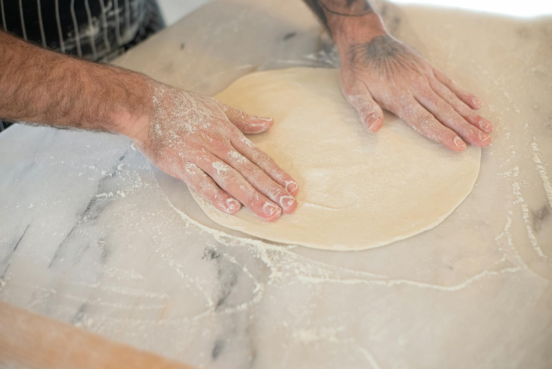 Pizza maker hands stretching fresh dough on flour-dusted marble surface in commercial kitchen